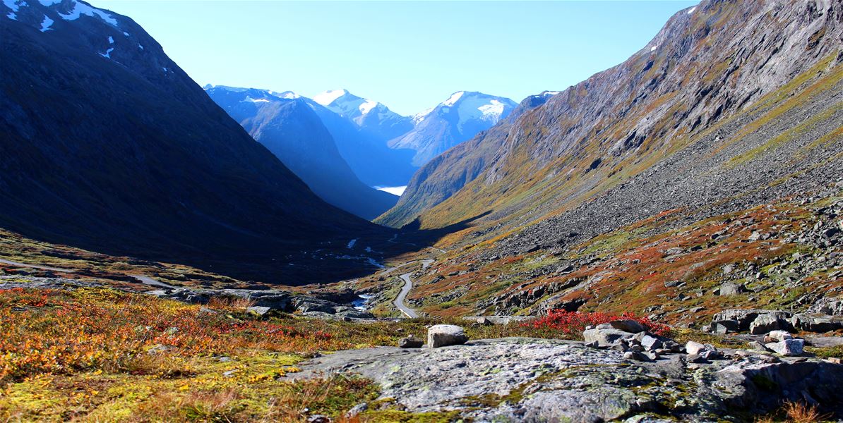 A winding road runs through a rocky mountain valley with autumn vegetation, steep slopes, and snow-capped peaks..