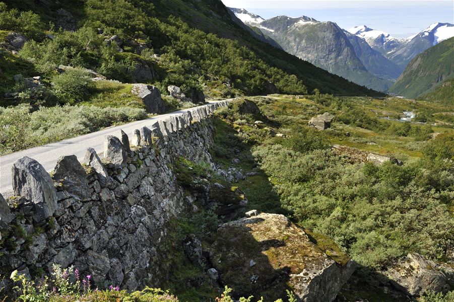 Narrow paved mountain road with a stone retaining wall, green shrubs, and distant snowcapped peaks in a valley..