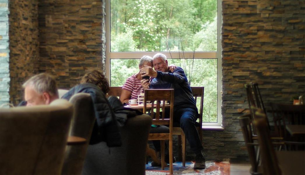 Older couple sits at a table by a large window, hugging while taking a selfie in a cozy cafe with stone walls..