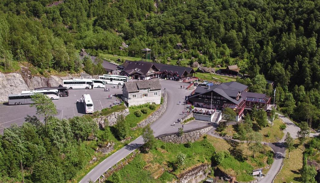 Aerial view of a mountain tourist stop with buildings, parked tour buses, and visitors surrounded by green forest..