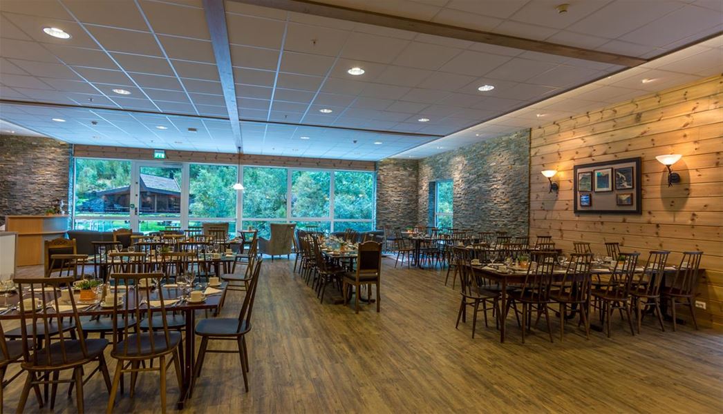 Empty rustic dining room with set tables and wooden chairs, stone and wood walls, and large windows facing trees..