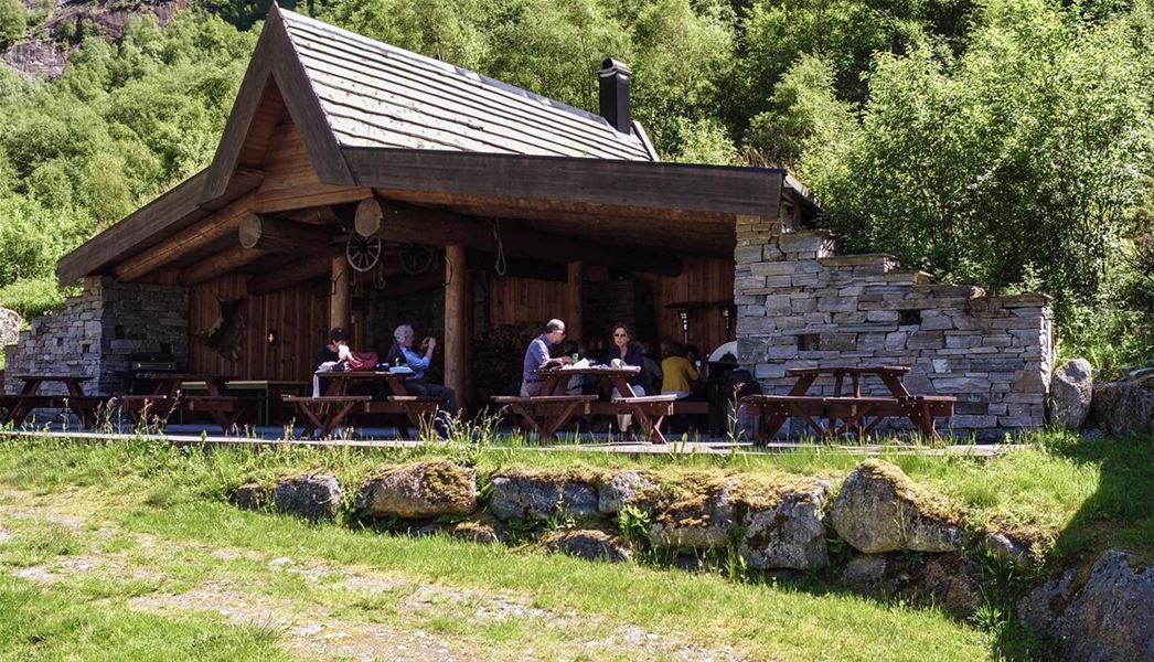 People sit at picnic tables outside a rustic wooden cabin with stone walls, surrounded by green trees in sunlight..