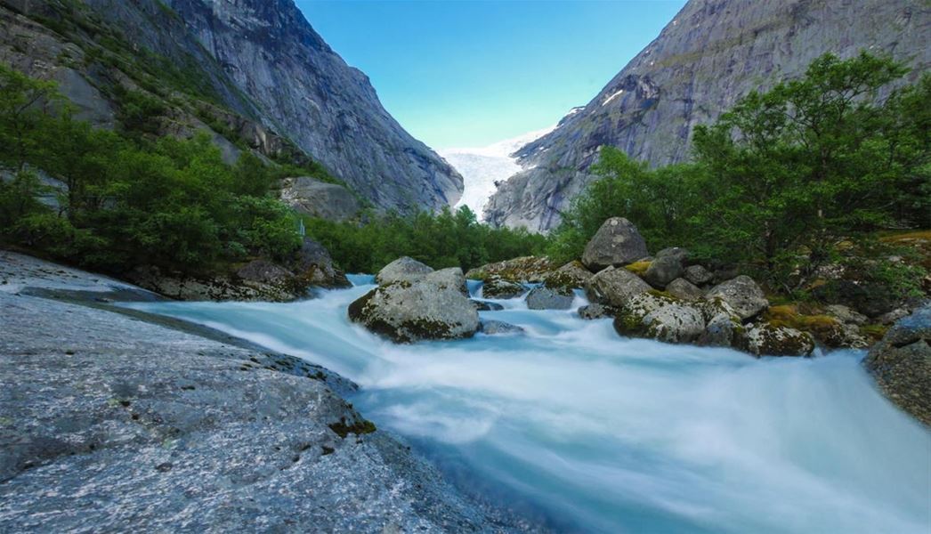 Fast-flowing milky blue river with boulders and green trees, framed by steep rocky mountains under blue sky..
