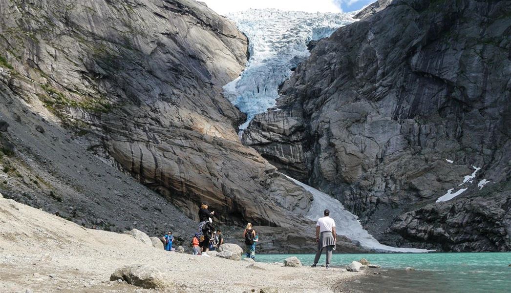 Hikers stand on a rocky shore beside a turquoise lake, facing a glacier descending between steep cliffs..