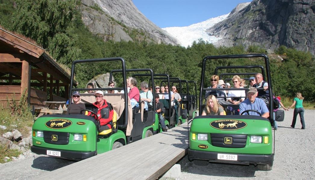 Group of tourists seated in green open shuttle carts on a gravel road with trees and a glacier between mountains..