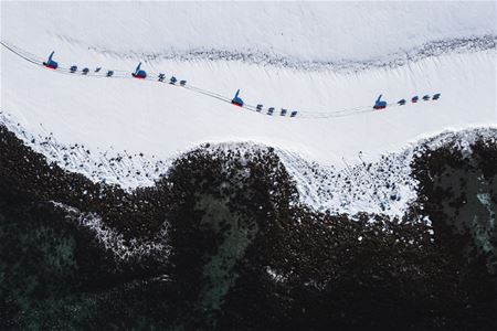 Hundespann kjører gjennom et snødekt arktisk landskap, sett ovenfra med mørke fjellformasjoner i forgrunnen.