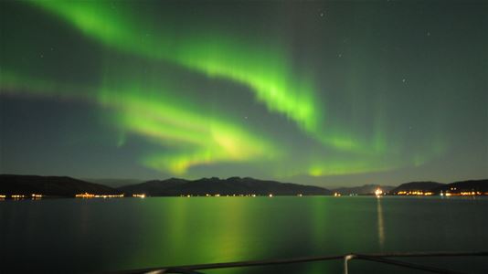 Grønt nordlys over fjorden og fjellene, sett fra en båt i Tromsø.