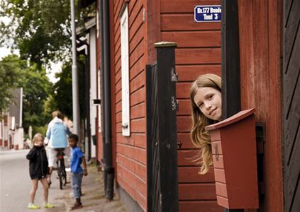 Rött äldre trähus, flicka som tittar fram vid en brevlåda, två barn på gatan och en kvinna som cyklar.