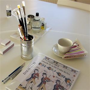 Ett bord dä det ligger en mapp med text dalamåleri, pennor, penslar i en mugg, en vit kaffekopp med en randig servett.