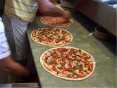 Tre pizzor på en bänk, man ser en del av en person i randig tröja och ett vitt förkläde som lägger på fyllning på pizzan.