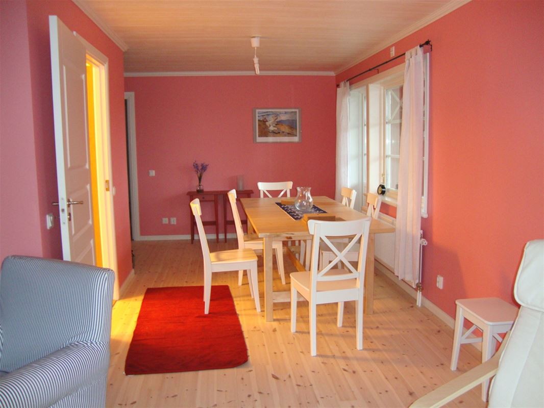 Rosa rum med röd matta och matbord med vita stolar framför ett fönster med vita gardiner. 