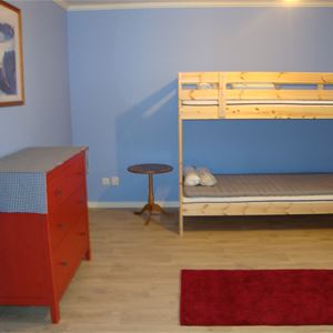 Blått rum med våningssäng av furu, röd byrå och röd matta på golvet.