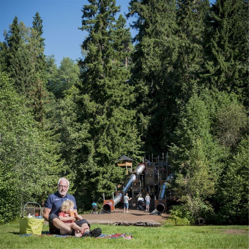 En man som sitter på en gräsmatta med en flicka i knät, grön picknickkorg brevid sig, rutschkanor i bakgrunden.