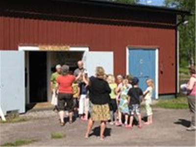 Barn och vuxna står samlade utanför en röd träbyggnad med öppna vita dörrar.