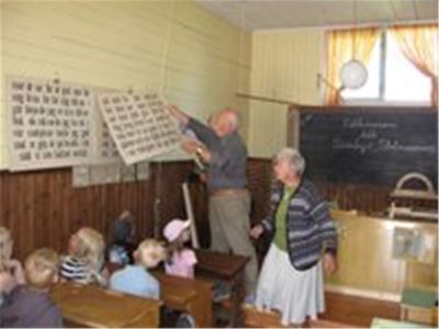 Children sitting in old school desks watching a man taking down a sign from the wall, a woman standing beside him.