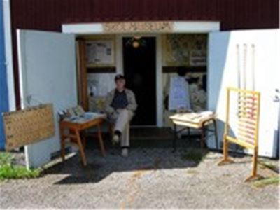 White open doors in the doorway sits a man in a black cap by an old school desk.