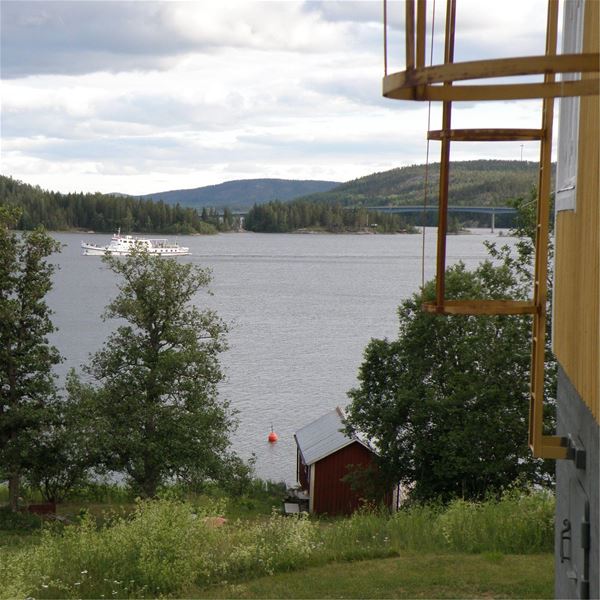Till höger på bilden syns kortsidan av huvudbyggnaden. Vid vattnet ligger sjöboden där det också finns logi. På fjärden passerar Ådalen III 