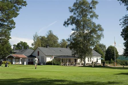 En gräsmatta, en person med en golfklubba, en vit byggnad med svart tak i bakgrunden.