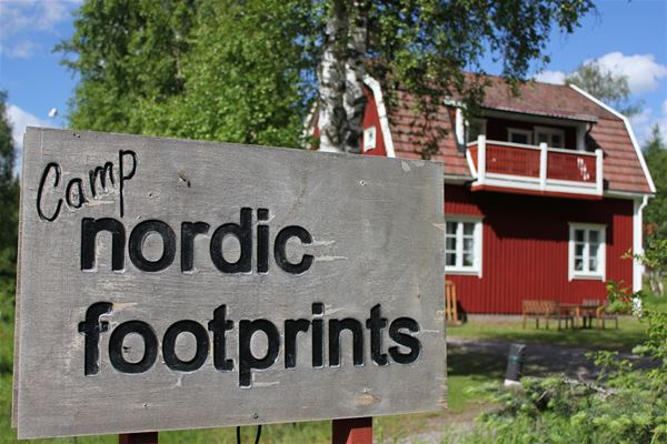 Nordic Footprints,  &copy; Nordic Footprints, Skylt med camp nordic footprints med ett rödmålat hus i bakgrunden. 
