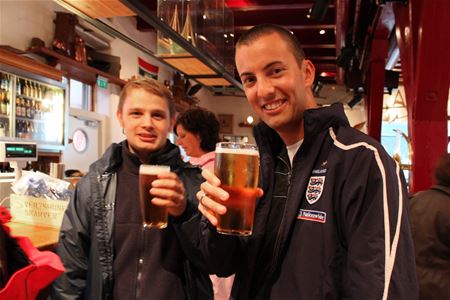 To personer skåler med øl i en bar, én iført England-jakke.