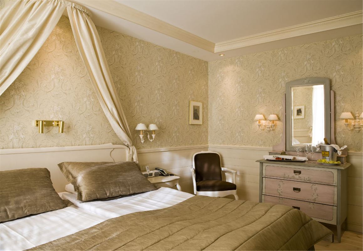 Hotel room with double bed, chest of drawers, mirror and a rococo chair.