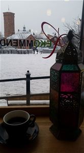 Wiew fom a window in the café, snow on the ground outside, a brick house with a tower, in the window a black cup and a lantern.