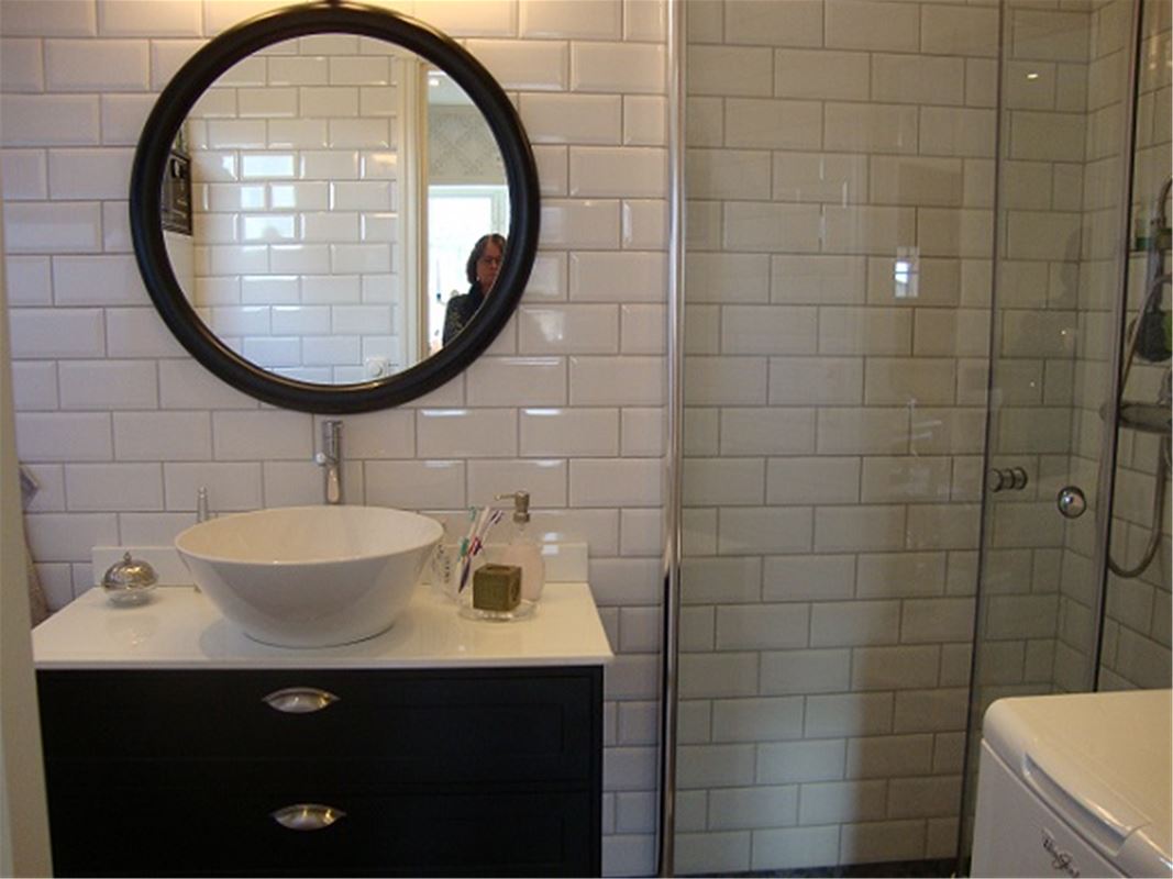Brun kommod med vitt handfat ovanpå, brun, rund spegel, duschväggar av glas och helkaklade, vita väggar.