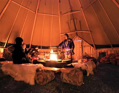 People gathered around a fire pit inside a large wooden tent structure with animal hides.