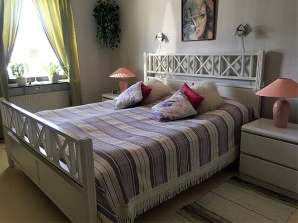 Dubbelsäng med vit sängram och lampor på vit byrå bredvid sängen.  