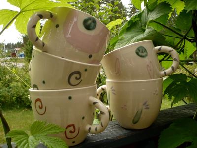 Fem vitglaserade keramikmuggar staplade på varandra, gröna blad runtomkring.