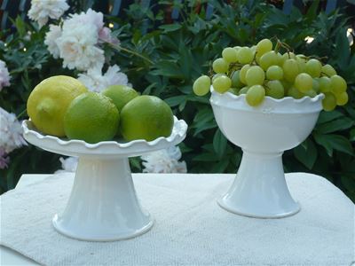 Två vitglaserade keramikfat på fot, i ett fat ligger gröna vindruvor, i det andra fyllt med gröna äpplen och en citron, faten står på en vit duk, vita blommor i bakgrunden.