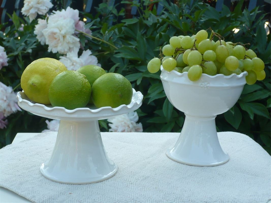 Two white glazed ceramic dishes on foot, on one dish are green grapes and on the other green apples and a lemon.