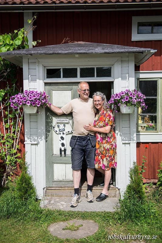 En man och en kvinna på en förstukvist, håller om varandra hängande lila petunior i krukor på var sida om paret.