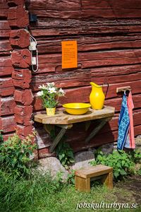 En rödmålad timmervägg, på ett bord som är fast i väggen står en gul zinkbalja och en gul kanna, blommor i en vas, två handdukar på varsin krok.