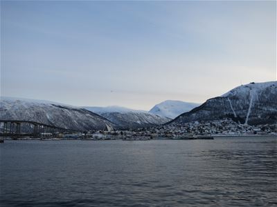 Tromsø with a bridge, buildings, and snow-covered mountains reflected in calm water.