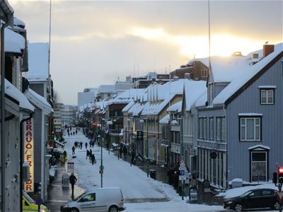 Snow-covered town street with people, cars, and buildings under a cloudy sky with a glimpse of sunlight.