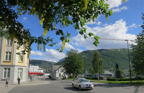 Street scene in Tromsø with people, a car, blooming trees, and mountains in the background.
