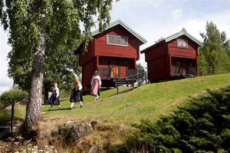 Three women in folklore costumes are walking on a lawn in front of two red timber cottages. 