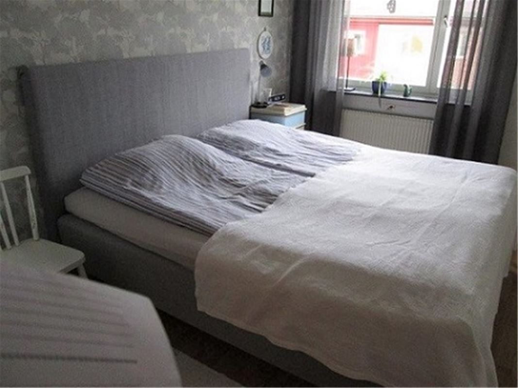 Dubbelsäng med ljusgrå sänggavel och grå, tunna gardiner i fönstret.