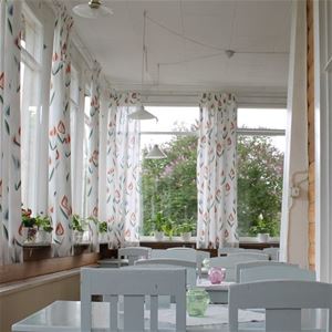 Verandafönstren med vita blommiga gardiner, blommor i fönstren och bord med stolar. 