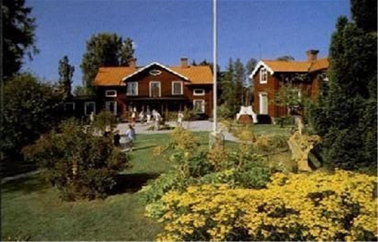 A garden, people sitting in the garden, two red wooden buildings in the background.