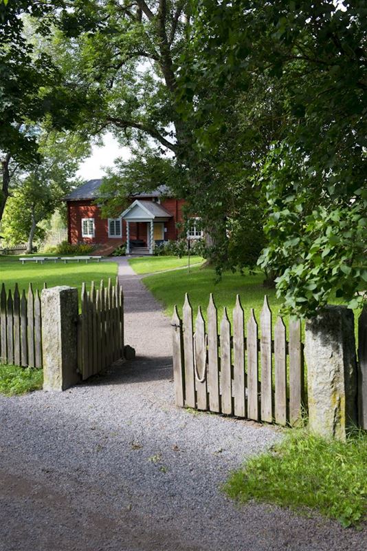 An open gate, a gravel walk, garden, a red timber building in two floors.