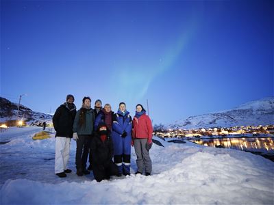 People under the Northern Lights in a snowy landscape with mountains and water in the background.