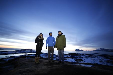 Three people on a snowy, rocky landscape at twilight with mountains and water in the background.