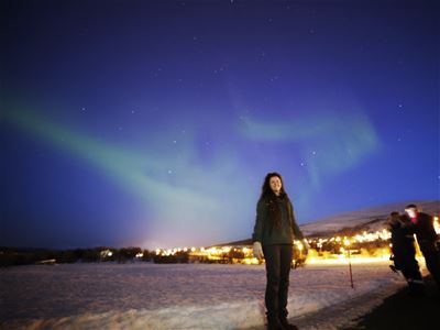 Person under the Northern Lights on a snowy plain with stars and buildings in the background.