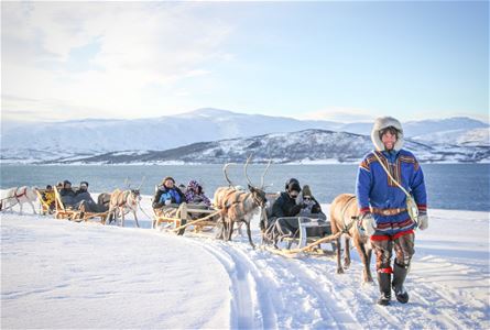 Reinsdyr trekker sleder med folk over snødekt landskap foran fjell og vann.