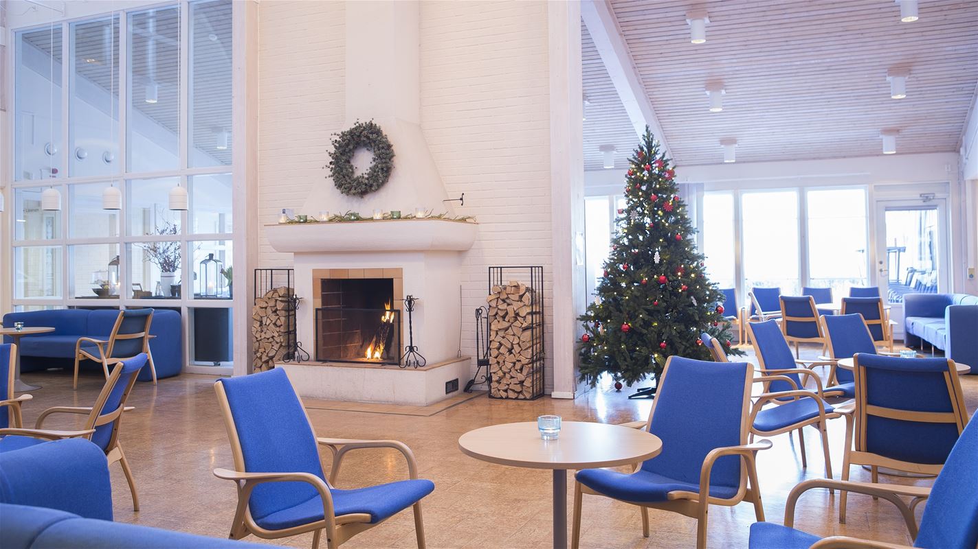 Large room with large windows and a large white fireplace in the middle of the room and several groups of blue bolstered chairs around tables in the room. 
