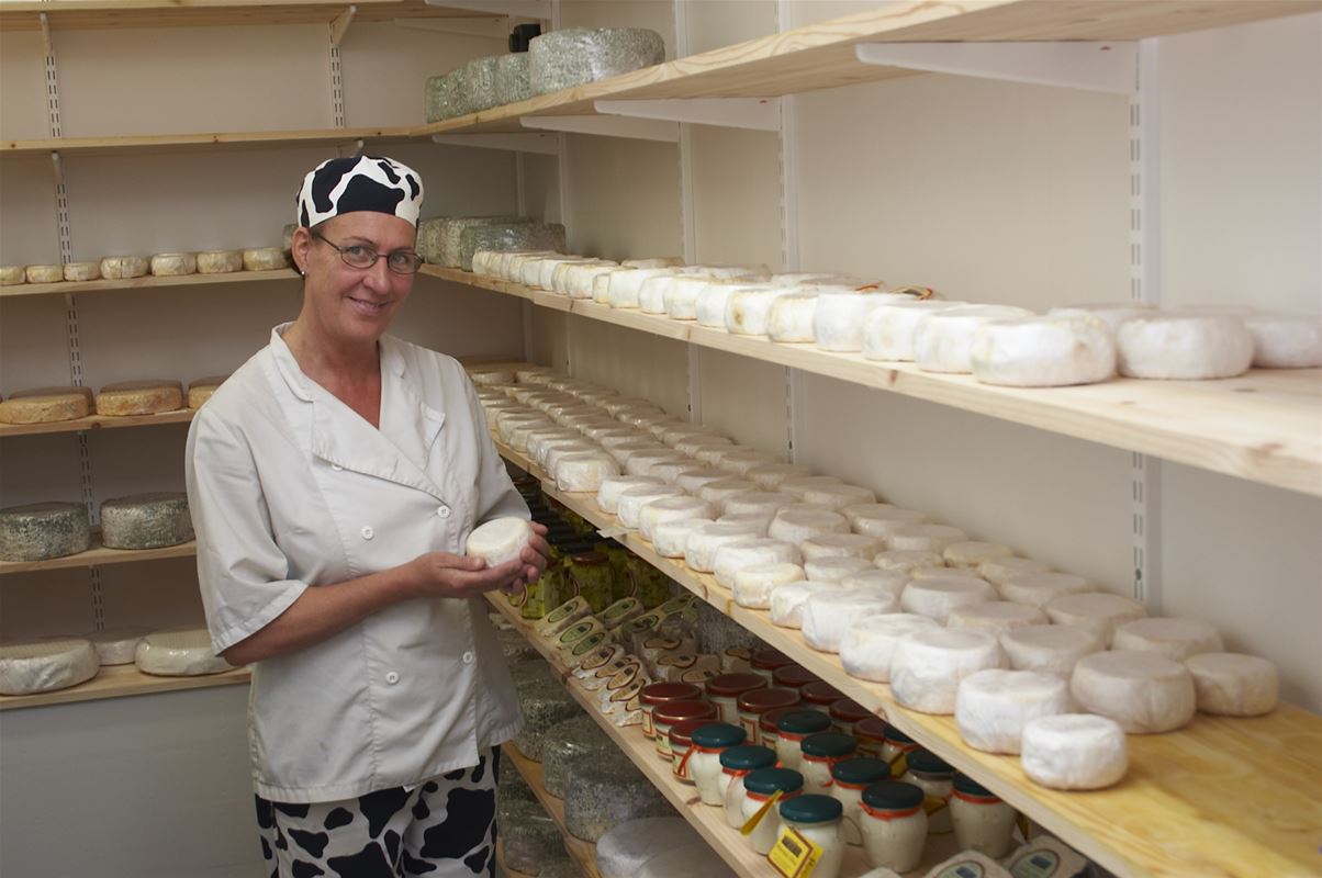 A woman in a white coat holding a cheese, cheeses lying on shelves around the room.