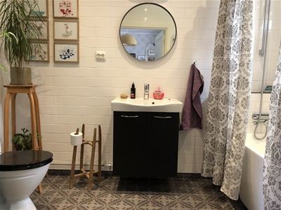 Ett badrum med vitt kakel, grått, mönstrat klinkers, toalett och en svart kommod med en rund spegel ovanför.