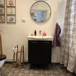 Bathroom with white tiled walls, grey, patterned floor and a black commode with a round mirror above.